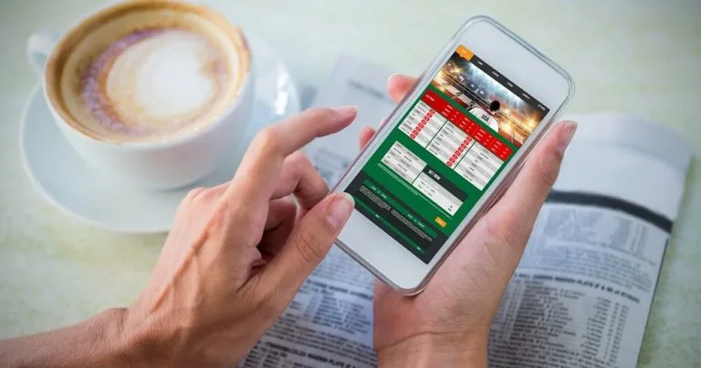 Manos sosteniendo un teléfono con una partida en pantalla junto a una taza de café con leche.