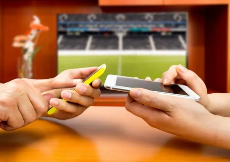 Dos manos sostienen un teléfono delante de una pantalla encendida con una cancha iluminada.
